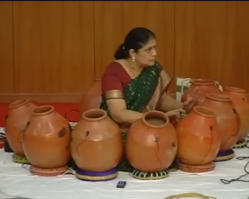Eine Frau steht vor einer ganzen Reihe von Ghatam-Instrumenten.